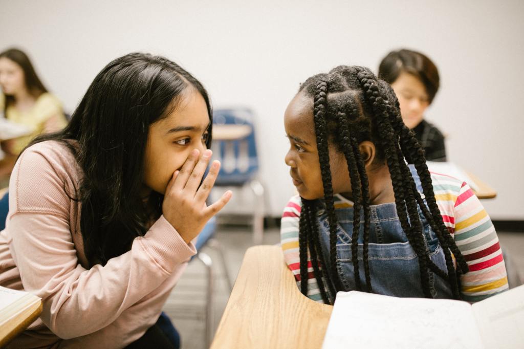 Two girls sit in a school classrom. One of them is whispering behind her hand to the other.