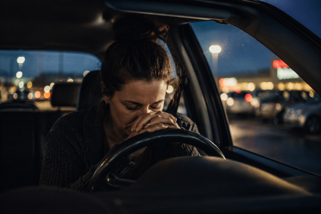 A tired woman sits alone in her car at dusk, resting against the steering wheel in a dimly lit parking lot, overwhelmed and emotionally drained after a stressful day.